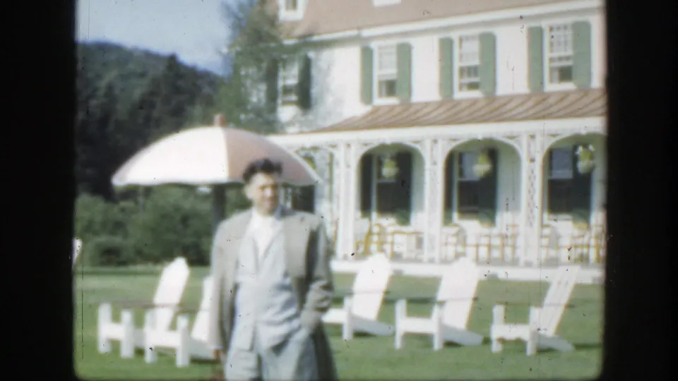 1950 Montreal frame with an adult man in a light suit on a green lawn beside white chairs, umbrellas, and a grand villa facade.