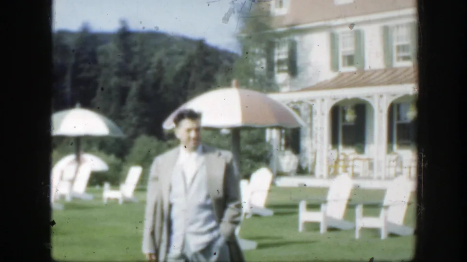 1950 Montreal home-movie still of a suited adult man on a resort-like lawn with white chairs, parasols, trees, and villa windows behind him.