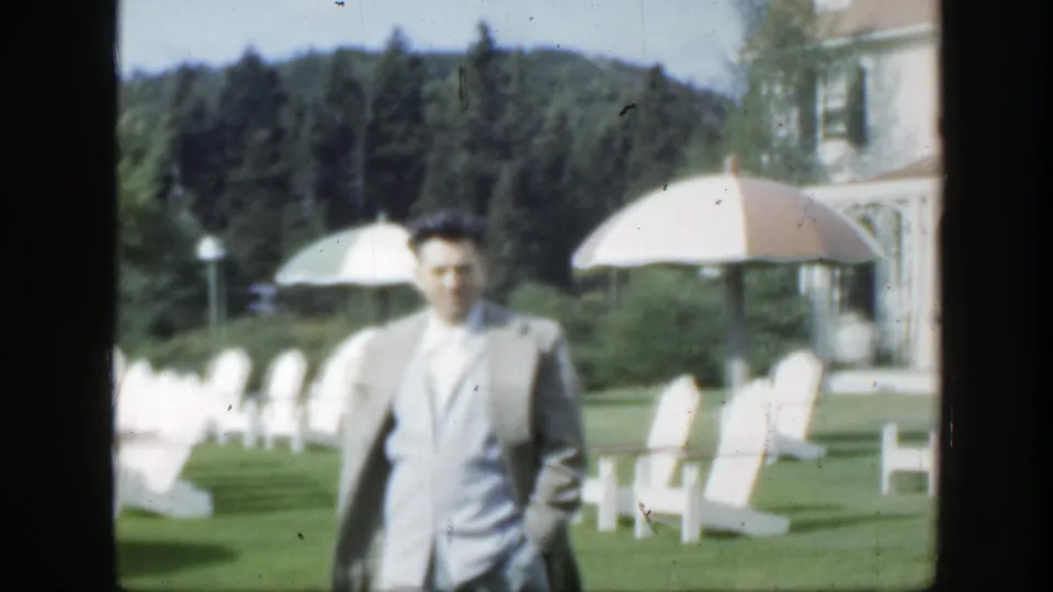 1950 Montreal frame showing the suited man closer to the camera, with white lawn chairs, parasols, trees, and part of the villa behind him.