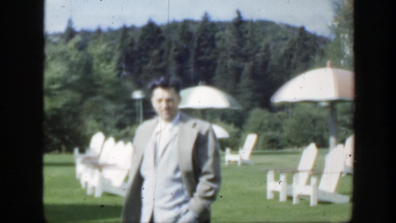 In 1950 Montreal home-movie footage, a man in a light suit approaches across a sunlit lawn with white chairs and umbrellas behind him.
