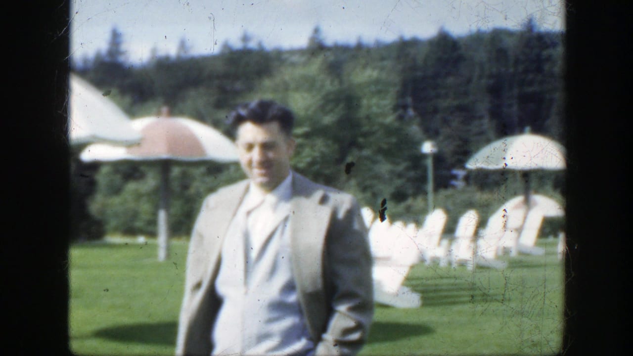 The suited man smiles while approaching the camera in a 1950 Montreal outdoor setting with lawn chairs, umbrellas and trees visible.