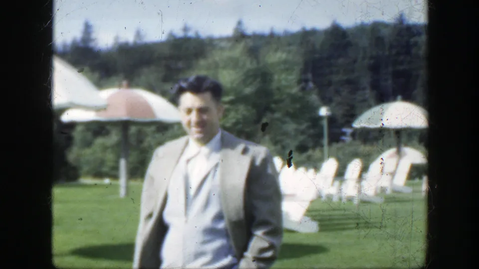 The suited man smiles while approaching the camera in a 1950 Montreal outdoor setting with lawn chairs, umbrellas and trees visible.