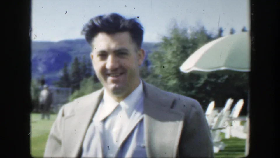 Close 1950 Montreal home-movie view of a smiling man in a light suit, with resort-style lawn chairs, umbrellas and hillside in the background.