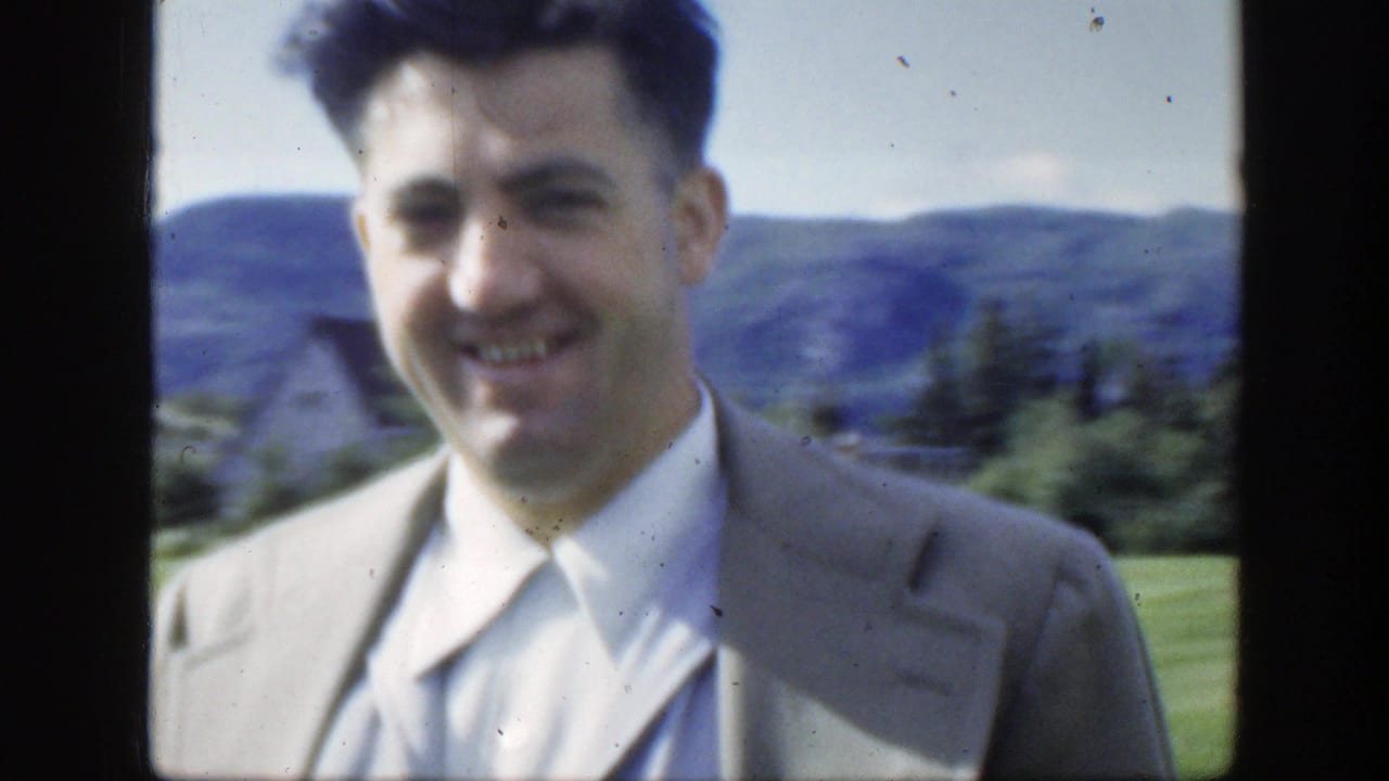 Man in a light suit smiles toward the camera with hills and lawn behind him.