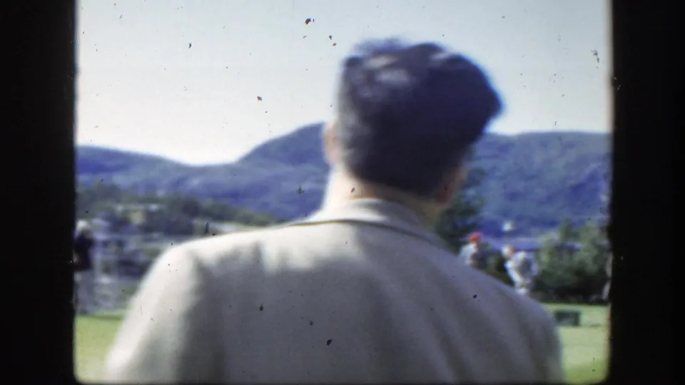 The man turns away from the camera on a grassy Montreal outdoor setting, with distant hills, trees, and other visitors visible.
