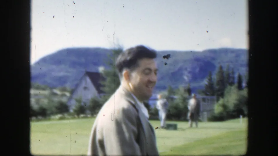 A 1950 Montreal home-movie frame shows the suited man laughing in profile on a green lawn as other adults stand farther back.