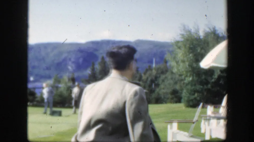 The suited man walks away across the grassy field in Montreal, with scattered people, trees, hills, and a pale umbrella at the edge of frame.