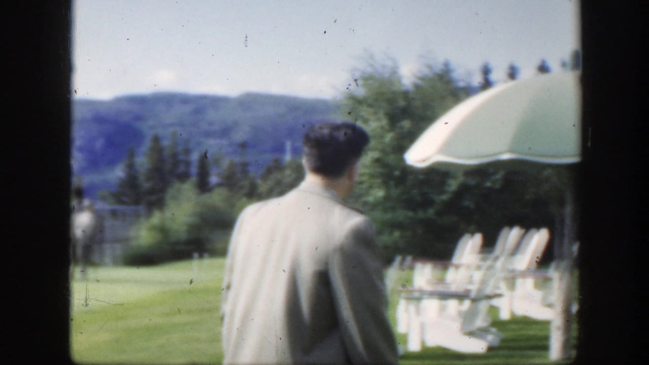 1950 Montreal home-movie frame of a man in a light jacket walking away across a lawn with white chairs, trees, and hills beyond.