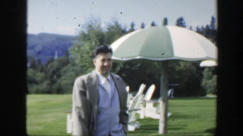 1950 Montreal home-movie still of a smiling adult man on a green lawn, with a striped parasol, white chairs, trees, and hills behind him.