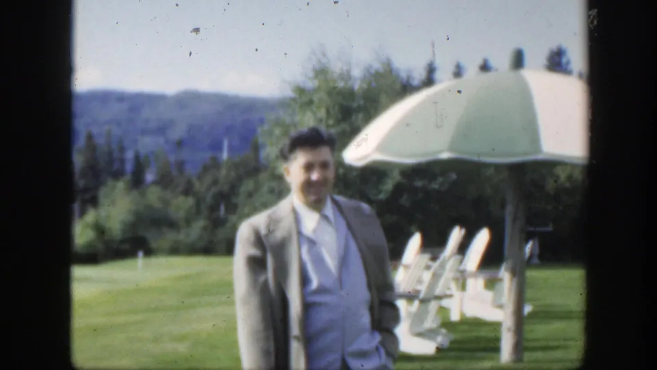 1950 Montreal frame of an adult man smiling on a lawn, with white garden chairs, a striped parasol, trees, and distant hills in view.