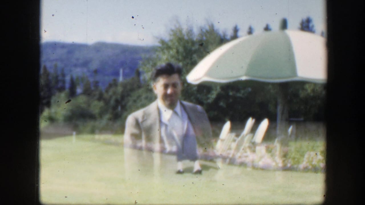 Man in a light suit standing on a lawn near a large umbrella with hills behind him.