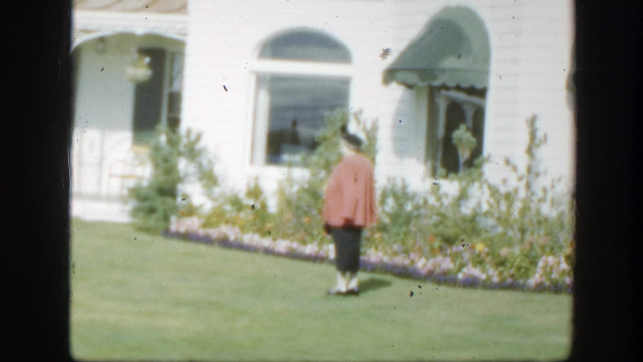 1950 Montreal home-movie frame of a woman walking across a green lawn toward a white house with arched windows, flowers, and green awnings.