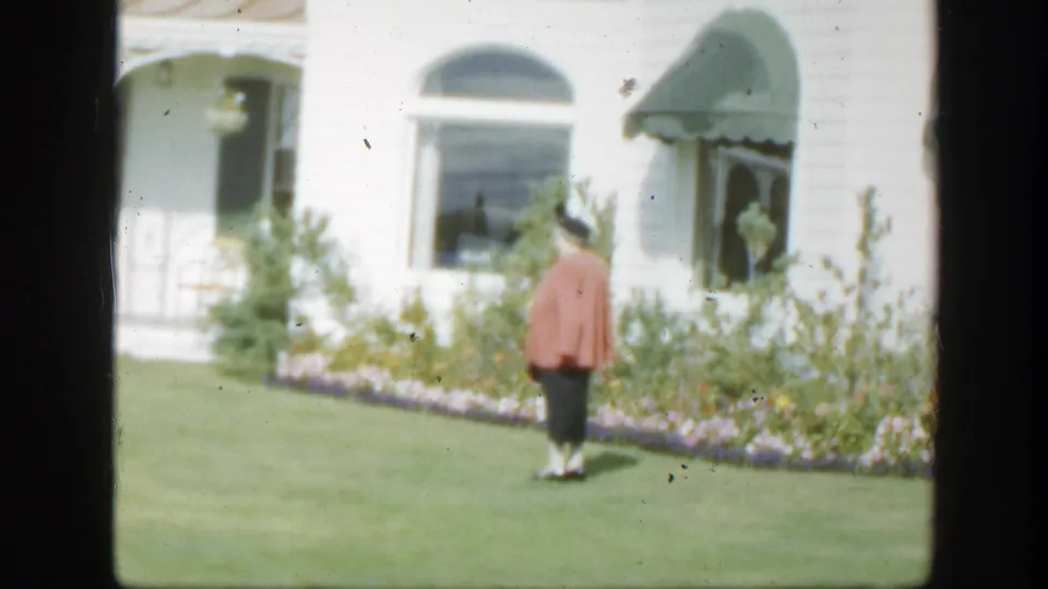 1950 Montreal home-movie frame of a woman walking across a green lawn toward a white house with arched windows, flowers, and green awnings.