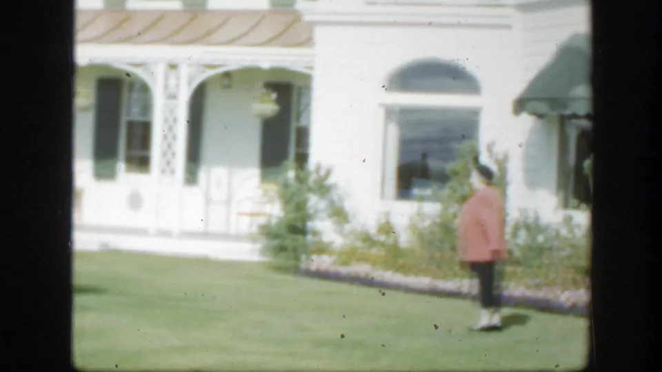 1950 Montreal home-movie frame showing a woman on a lawn beside a white house with decorative porch arches, flower beds, and green shutters or awnings.