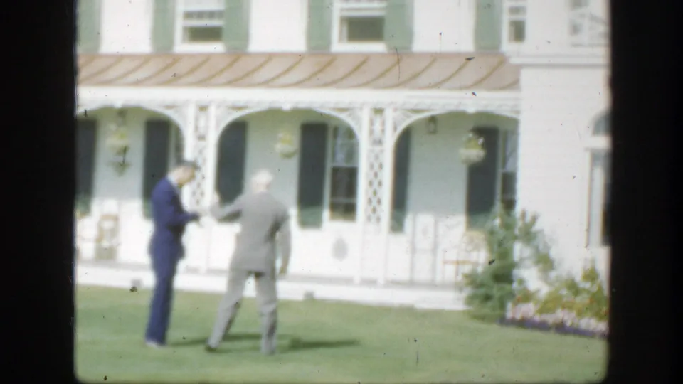 1950 Montreal home-movie frame of two suited men conversing on the lawn in front of a white house porch with arched supports and hanging planters.