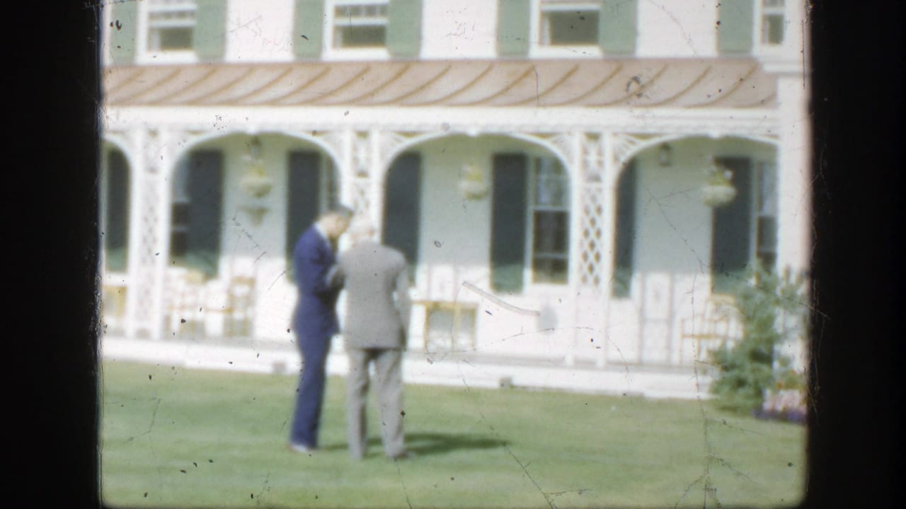 In 1950 Montreal home-movie footage, two suited men converse on a manicured lawn before a white house with green shutters.