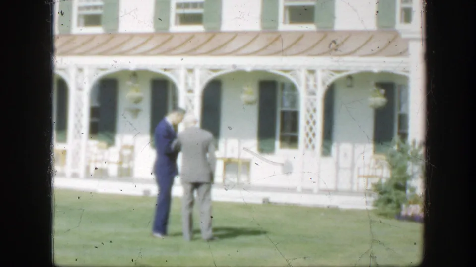 In 1950 Montreal home-movie footage, two suited men converse on a manicured lawn before a white house with green shutters.