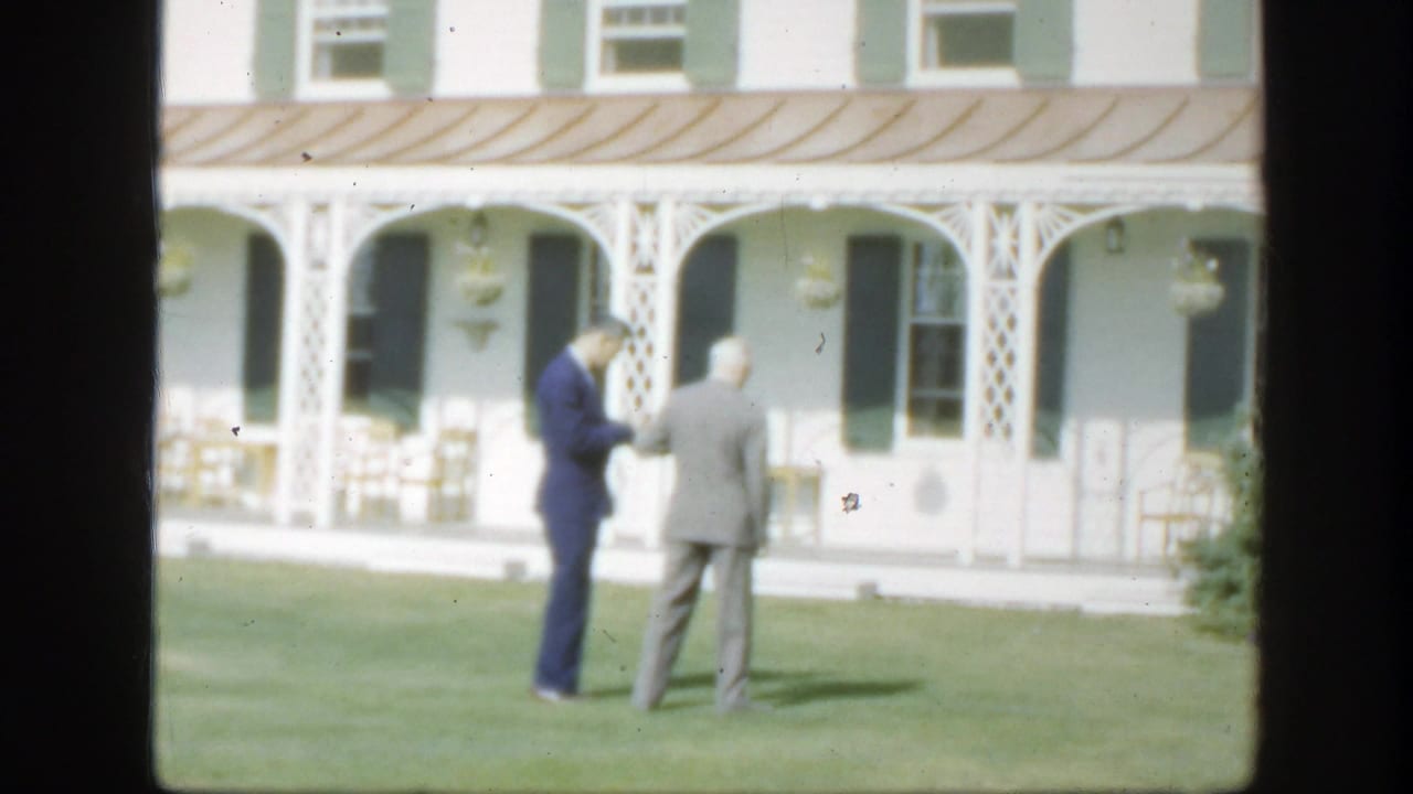 Two suited men continue their lawn-side conversation outside a white Montreal house with green shutters and porch columns.