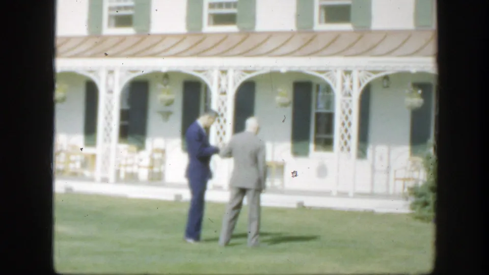 Two suited men continue their lawn-side conversation outside a white Montreal house with green shutters and porch columns.