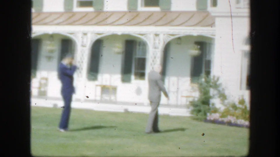 One suited man stands near the left of frame as another walks away across the lawn outside the white house.