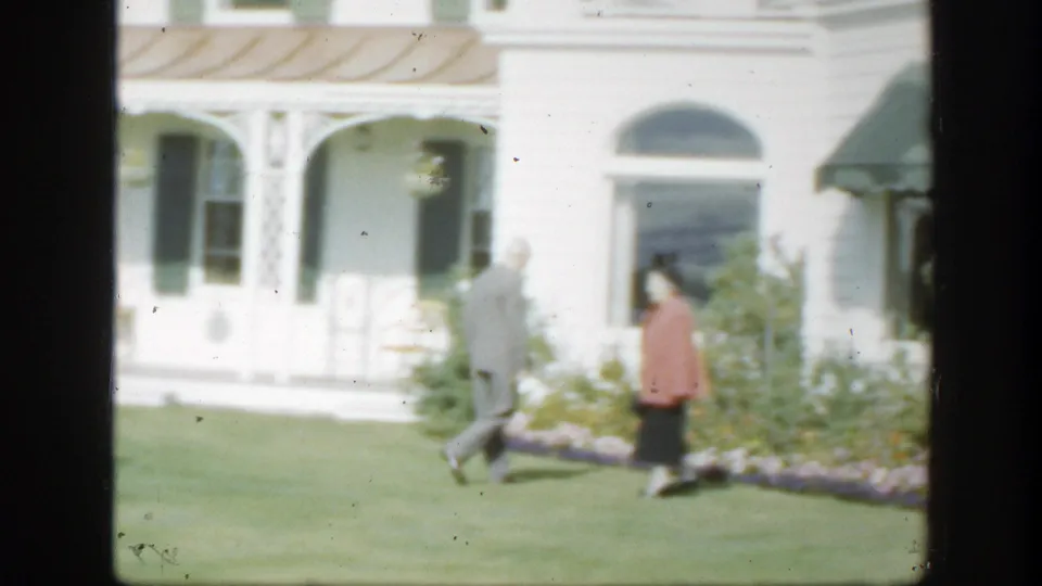 A woman in a pink top and dark skirt enters the 1950 Montreal lawn scene as a suited man walks toward the garden.