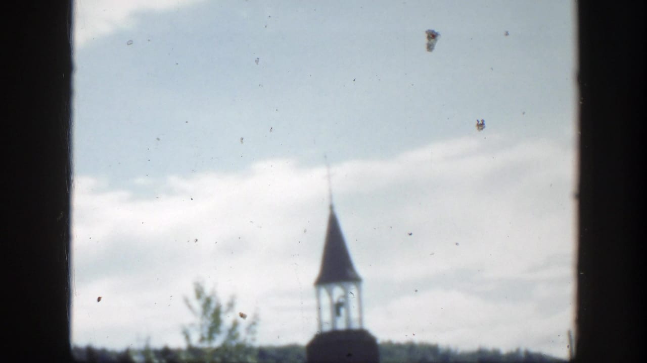 1950 Montreal home-movie frame showing a church steeple partly obscured by trees, with hills and cloudy sky behind it.