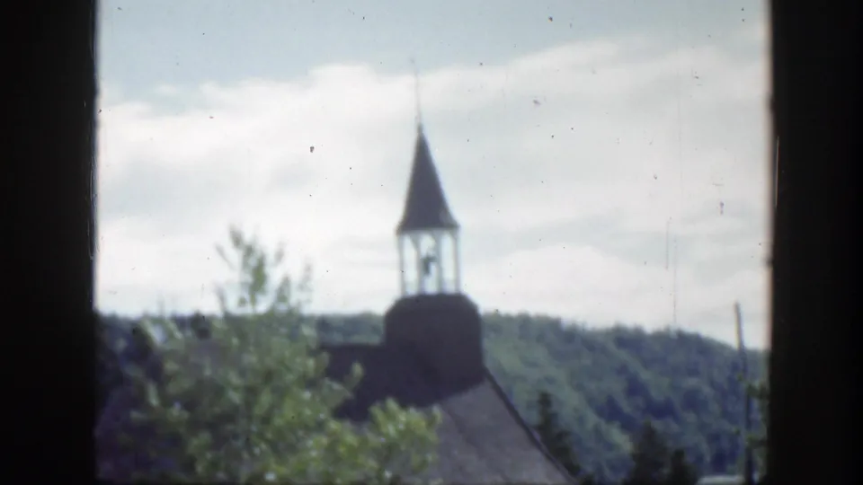 1950 Montreal frame of a small white church with steeple, partly hidden by leafy trees and backed by wooded hills.