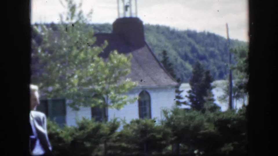 1950 Montreal home-movie view of a white church and lawn, with trees in front and suited adults partly entering the frame.