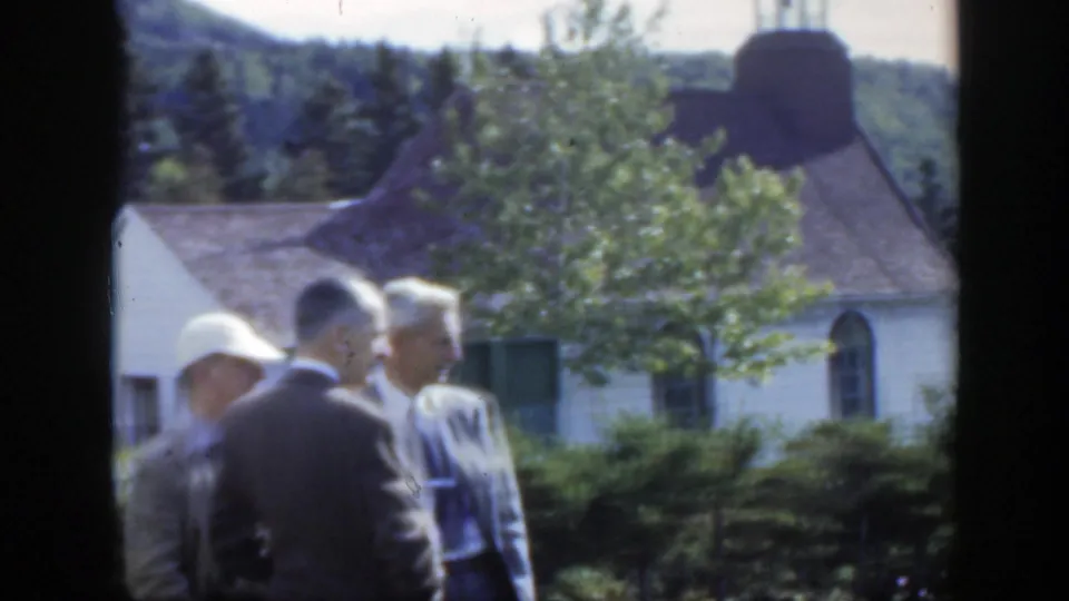 1950 Montreal frame showing three suited men conversing on a lawn outside a small white church with trees and hills behind them.