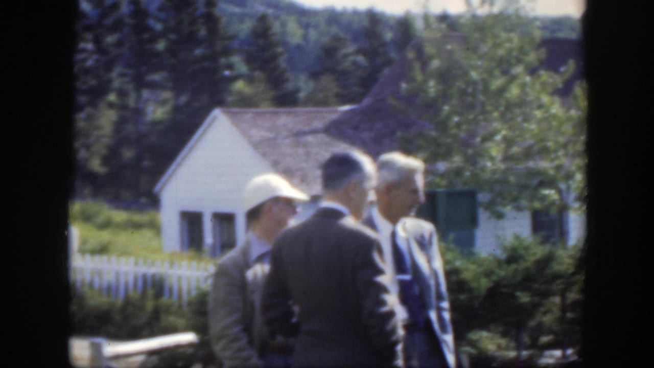 In 1950 Montreal-area home-movie footage, men in suits stand near a fenced rural cemetery with a small white building and trees behind them.