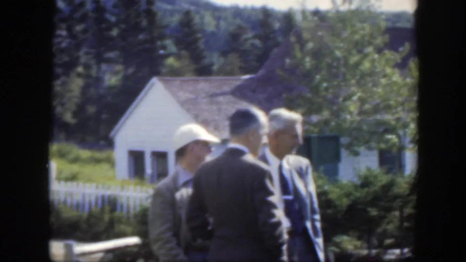 In 1950 Montreal-area home-movie footage, men in suits stand near a fenced rural cemetery with a small white building and trees behind them.