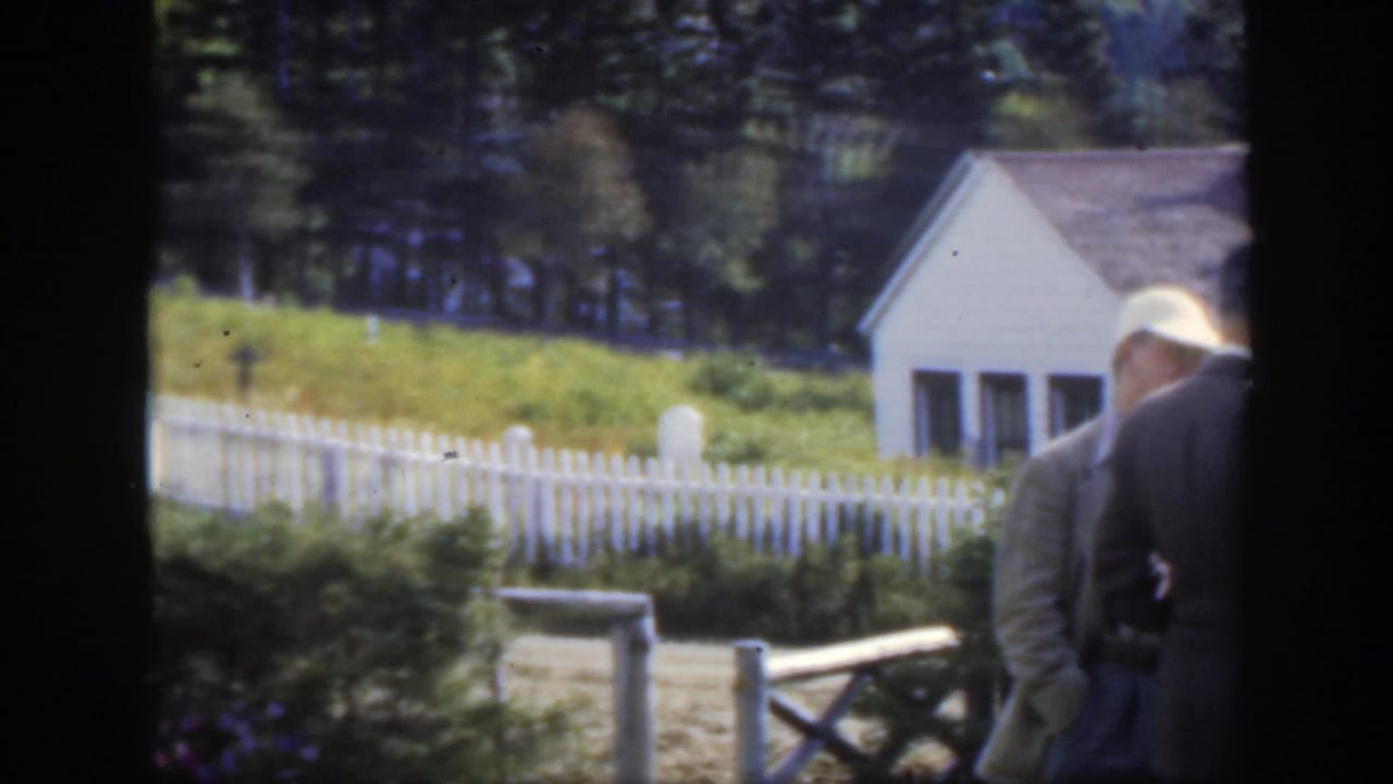 The camera looks across a white picket fence toward grave markers, grass, trees, and a small white building in the Montreal-area cemetery scene.
