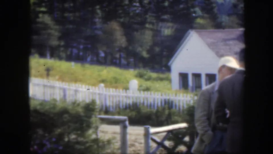 The camera looks across a white picket fence toward grave markers, grass, trees, and a small white building in the Montreal-area cemetery scene.