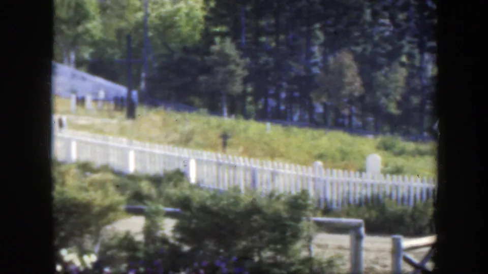 The 8mm view holds on the fenced cemetery landscape, showing pale grave markers, grass, trees, and a small building at the edge of frame.