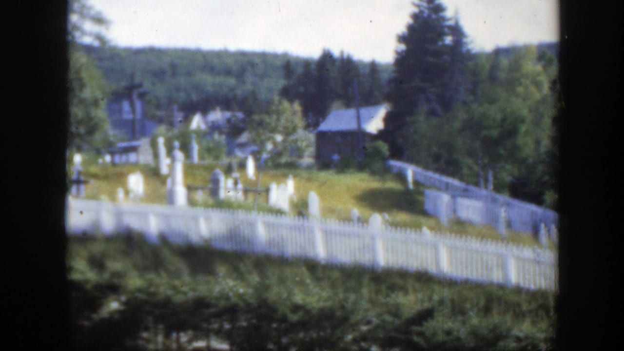 1950 Montreal-area 8mm view of a white fenced cemetery with grave markers, trees, small buildings, and a distant wooded hill.