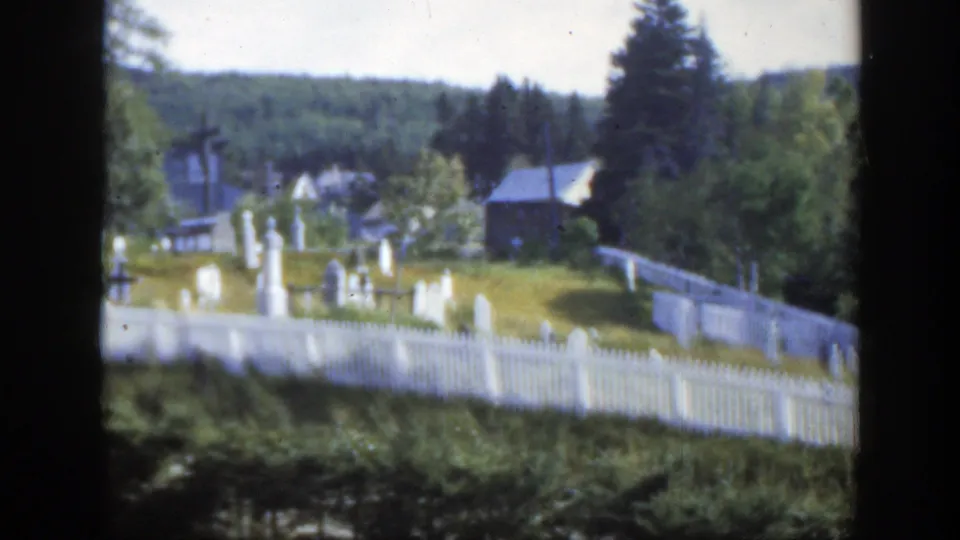 1950 Montreal-area 8mm view of a white fenced cemetery with grave markers, trees, small buildings, and a distant wooded hill.