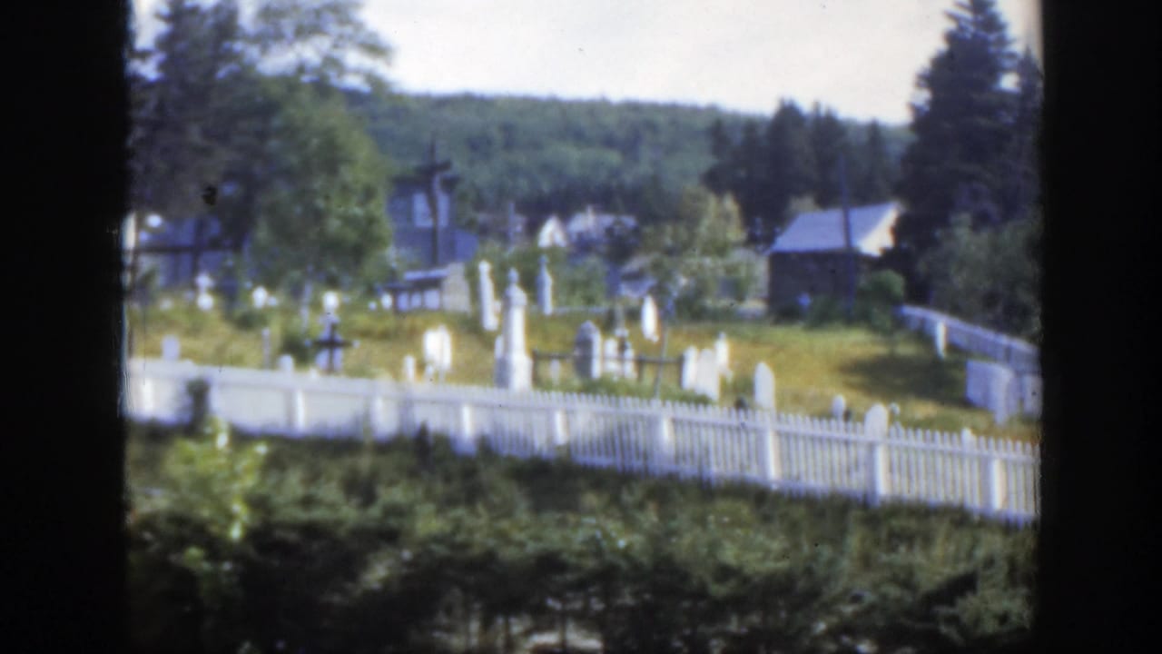 Cemetery grounds with white fence, gravestones, trees, buildings, and forested hill behind.