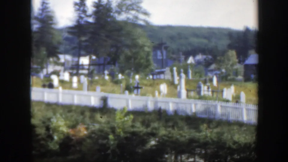 1950 Montreal-area 8mm frame looking across a white cemetery fence toward headstones, a cross, trees, buildings, and a green hillside.