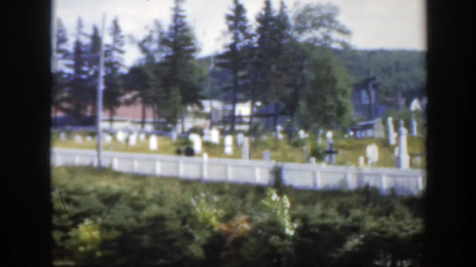 1950 Montreal-area home-movie view of a white fenced cemetery with headstones, a cross, trees, a utility pole, buildings, and wooded hillside.