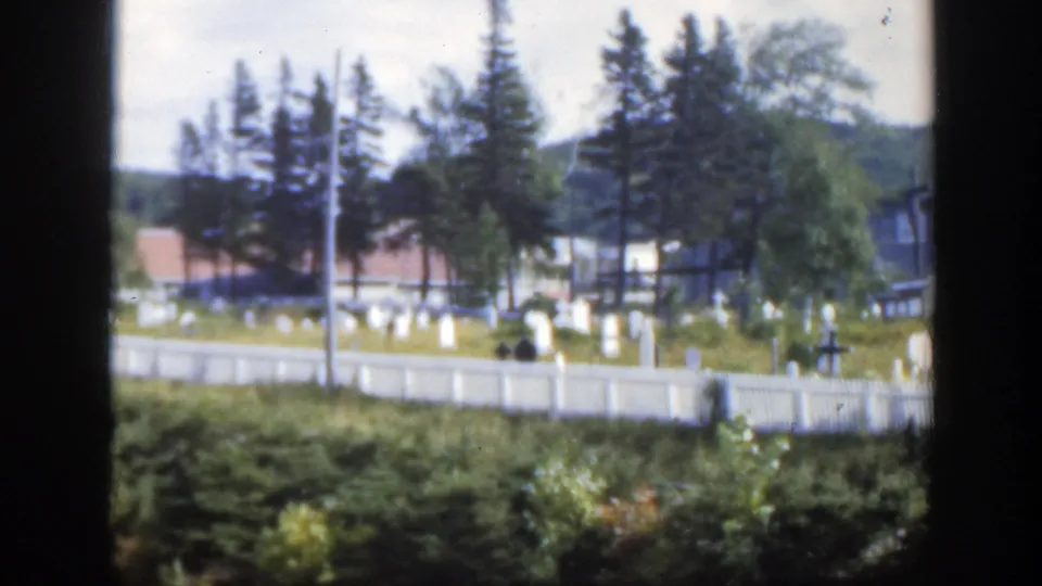 1950 Montreal cemetery view with pale headstones, a white fence, evergreens, and red-roofed buildings seen across foreground greenery.