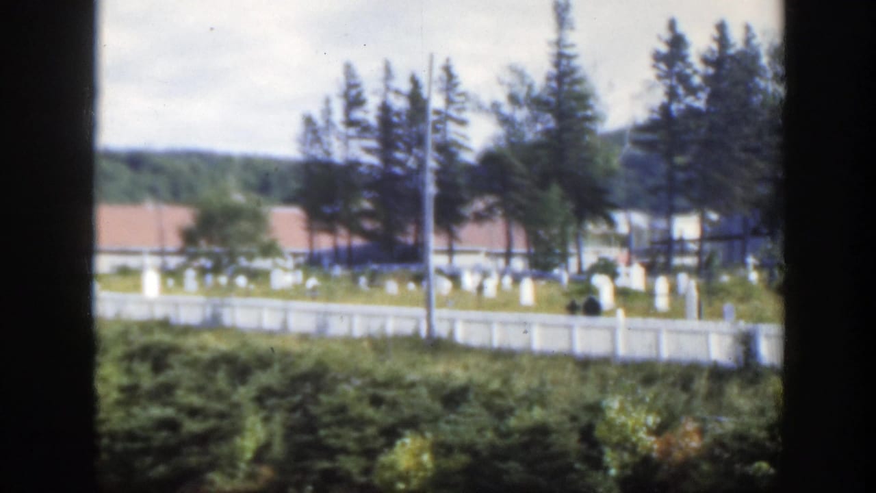 Cemetery grounds with headstones, white fence, trees, red roofs, and distant hills.