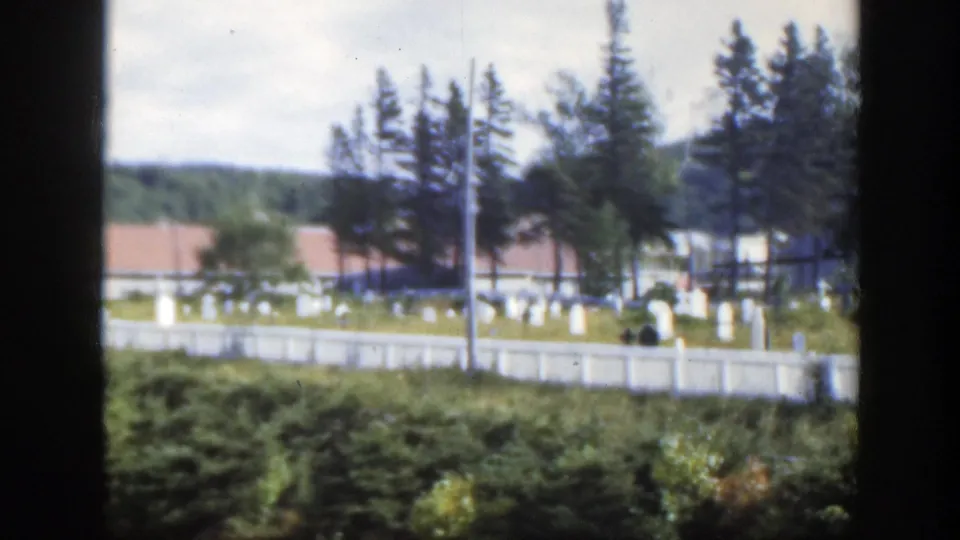 Cemetery grounds in Montreal with rows of pale headstones, a white fence, evergreens, red-roofed structures, and low wooded hills.