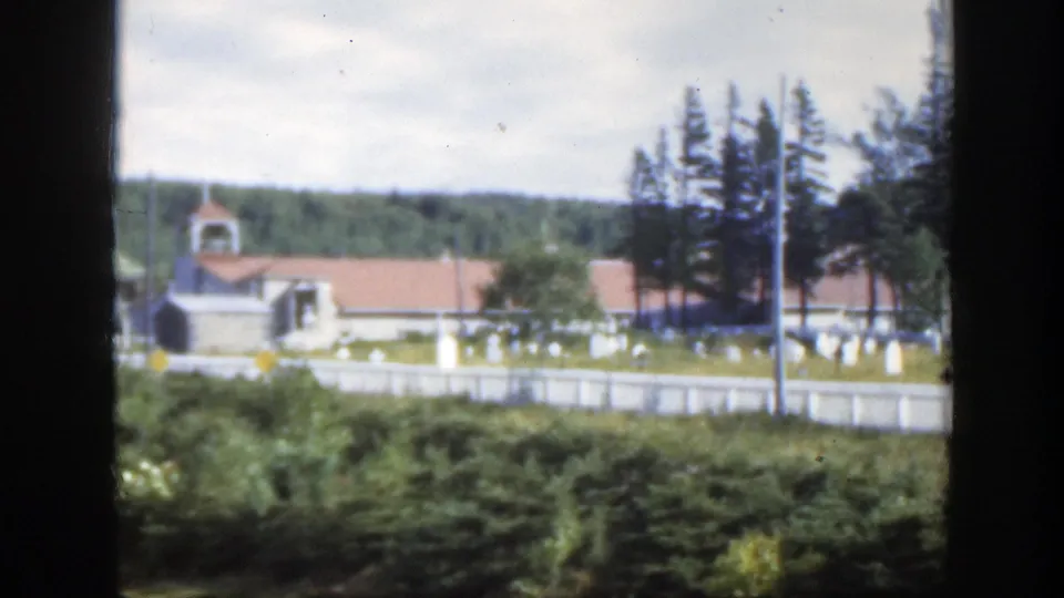 1950 Montreal cemetery scene with a small steepled church, pale headstones, white fence, evergreens, and dense foreground greenery.