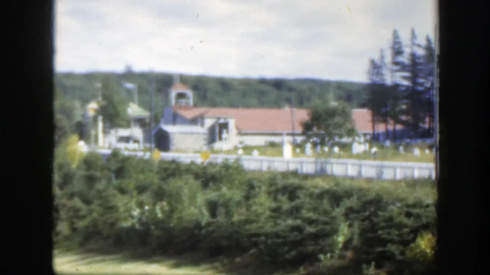 Montreal cemetery and church view showing a steepled building, white headstones, a low fence, evergreens, shrubs, and wooded hills.