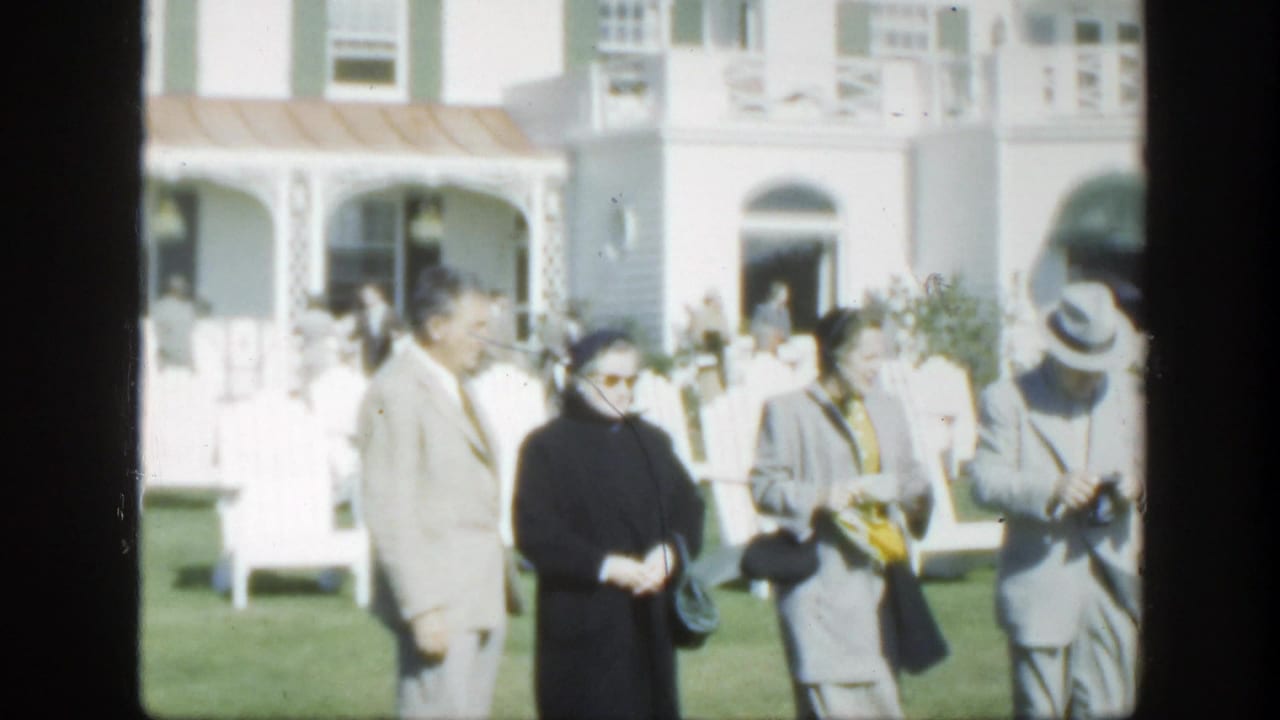 Older adults standing on a lawn outside a white building with arched openings.