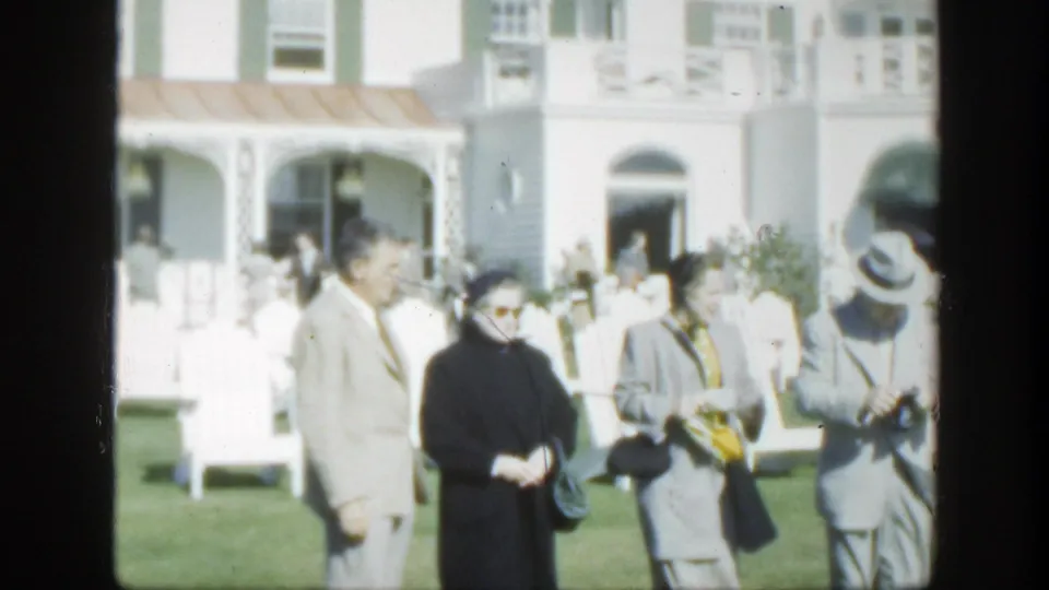 Older adults in mid-century coats and hats stand on a lawn outside a white building in Montreal, with arched openings behind them.