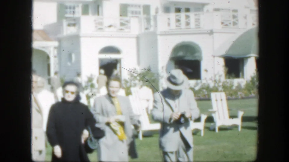 1950 Montreal frame showing adults on a lawn, one holding a camera, with white chairs and a green awning near the building.