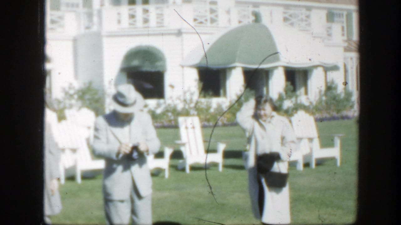 1950 Montreal home-movie frame showing two adults with cameras on a sunny lawn before a white house with green awnings.
