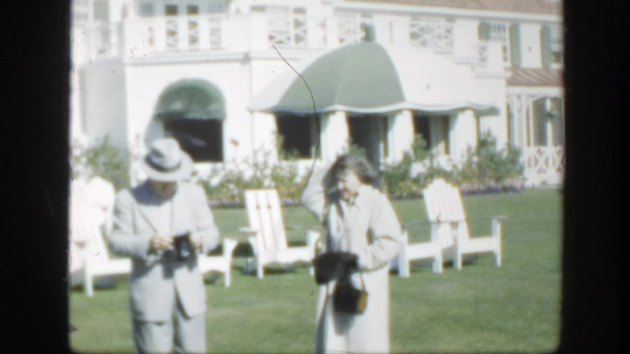 1950 Montreal frame of a man holding a camera beside a woman in a light coat, with white chairs and a house behind them.