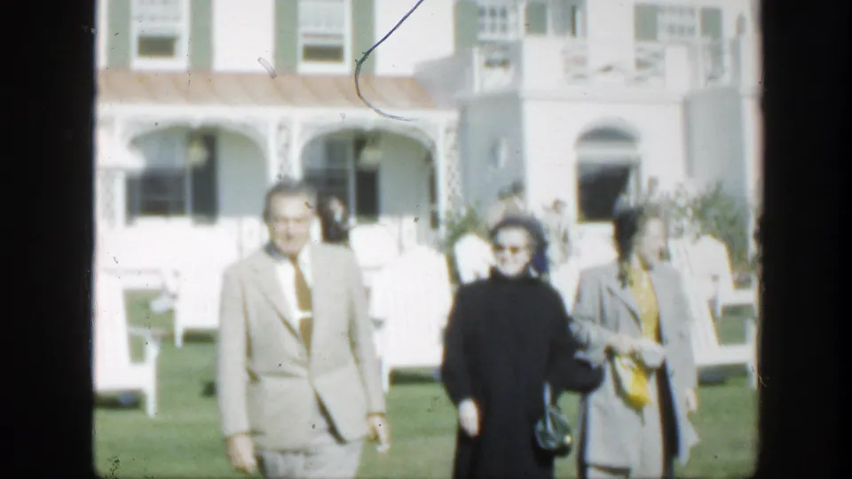 1950 Montreal home-movie frame showing three adults crossing a bright lawn before a white residence with porch railings.